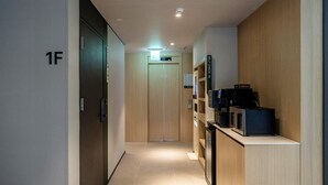 Un pasillo de hotel con suelo de madera, una recepción con una máquina de café y una puerta con la etiqueta '1F'.