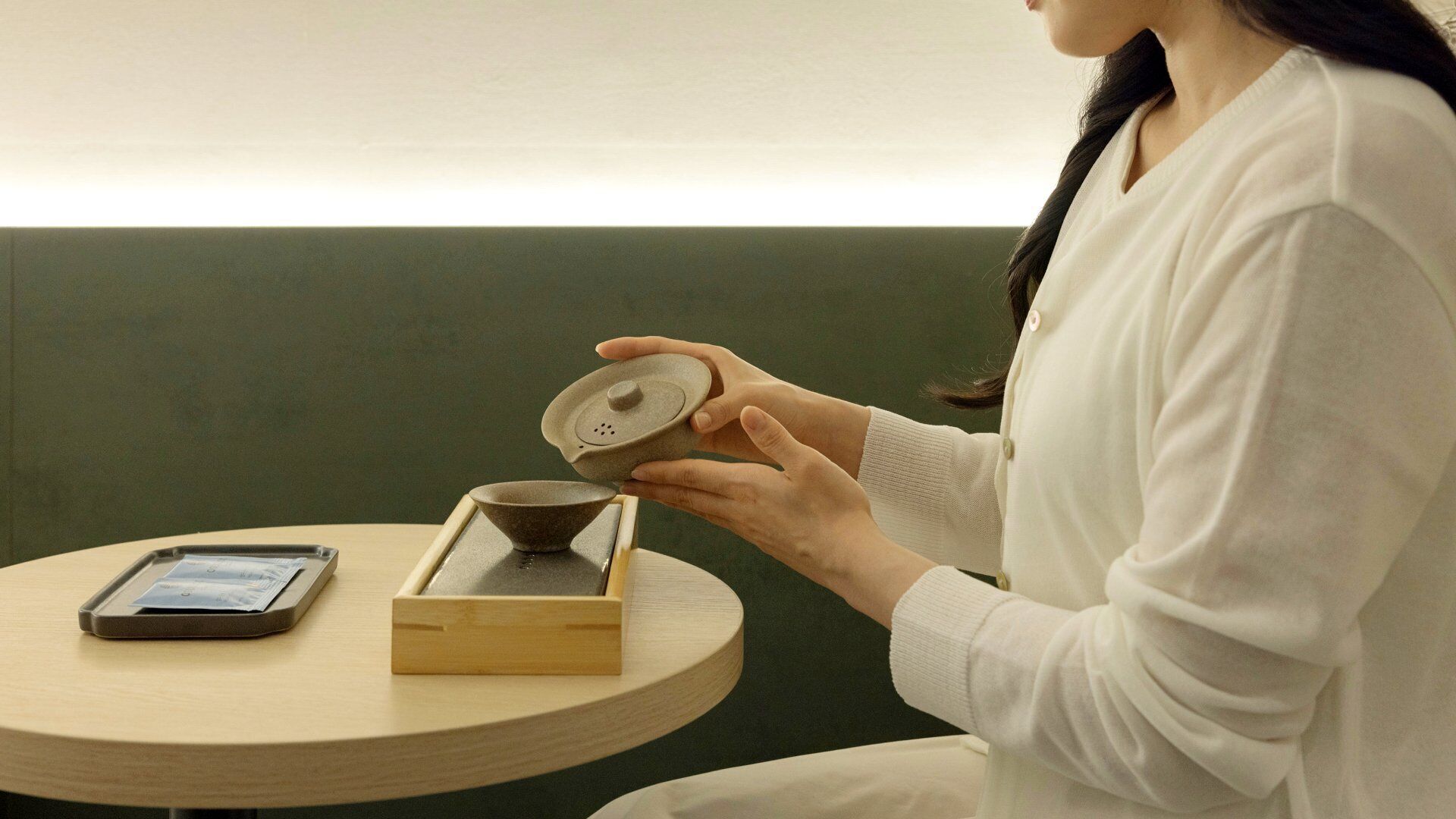 Una persona sosteniendo un cuenco de cerámica, sentada a una mesa redonda de madera con un teléfono inteligente y una caja de té.