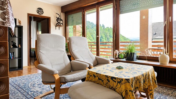 Ein Zimmer mit einem großen Fenster, einem Tisch mit einem gemusterten Tischläufer und einem Bücherregal.