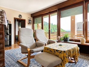 Una habitación con un ventanal grande, una mesa con mantel estampado y una estantería.