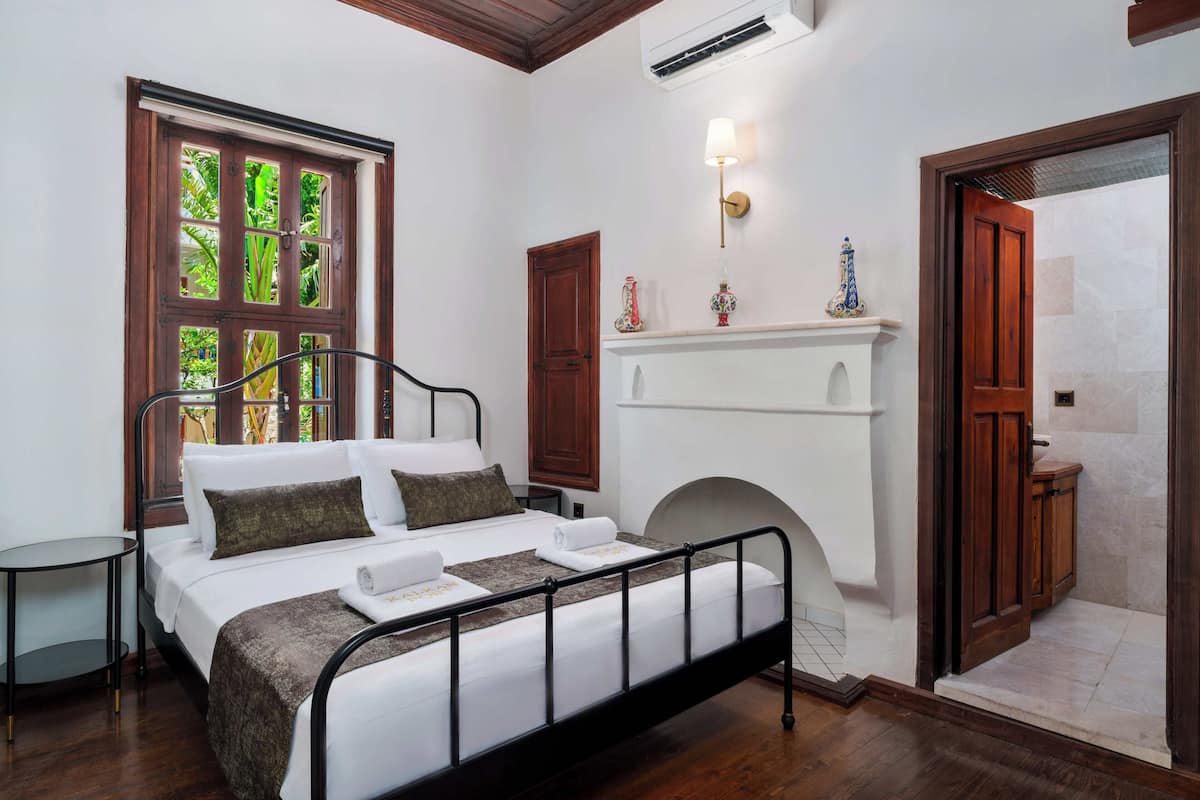 Quarto com cama, mesa de cabeceira, janela com vista, lareira e porta para outro cômodo.
