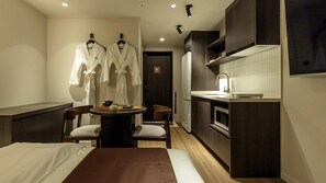 Habitación de hotel compacta con cama, una pequeña mesa de comedor, dos sillas, kitchenette y dos batas colgadas en la pared.