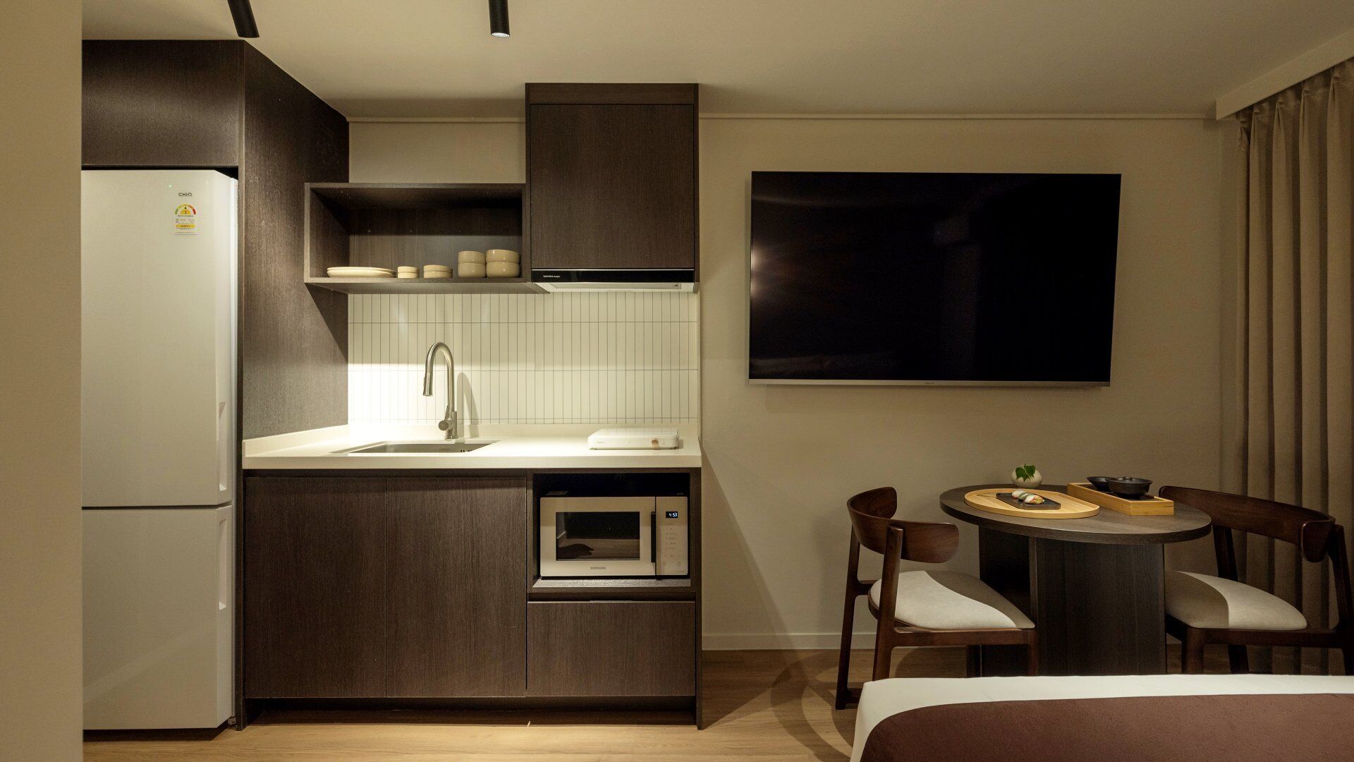 Une cuisine compacte équipée d’un réfrigérateur, d’un four à micro-ondes et d’un évier, un coin repas avec une table et des chaises, et un grand téléviseur à écran plat fixé au mur.