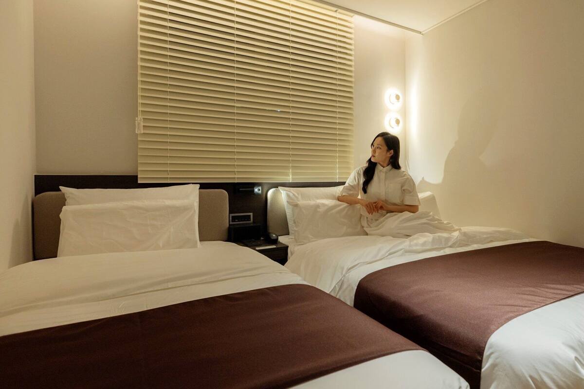 Habitación de hotel con dos camas, una mesita de noche, una lámpara de pared y un ventanal con persianas.