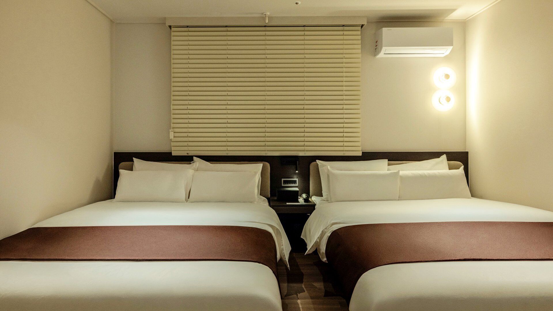 A hotel room with two beds, a window with blinds, an air conditioning unit, and a bedside table with a clock.