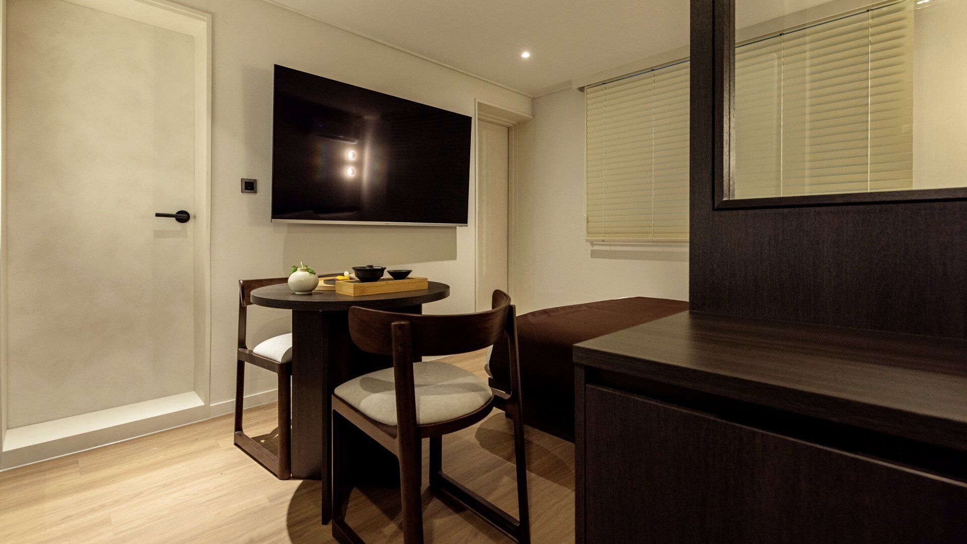 A compact room with a flat-screen TV mounted on the wall, a small round table with chairs, a wooden cabinet, and a window with blinds.