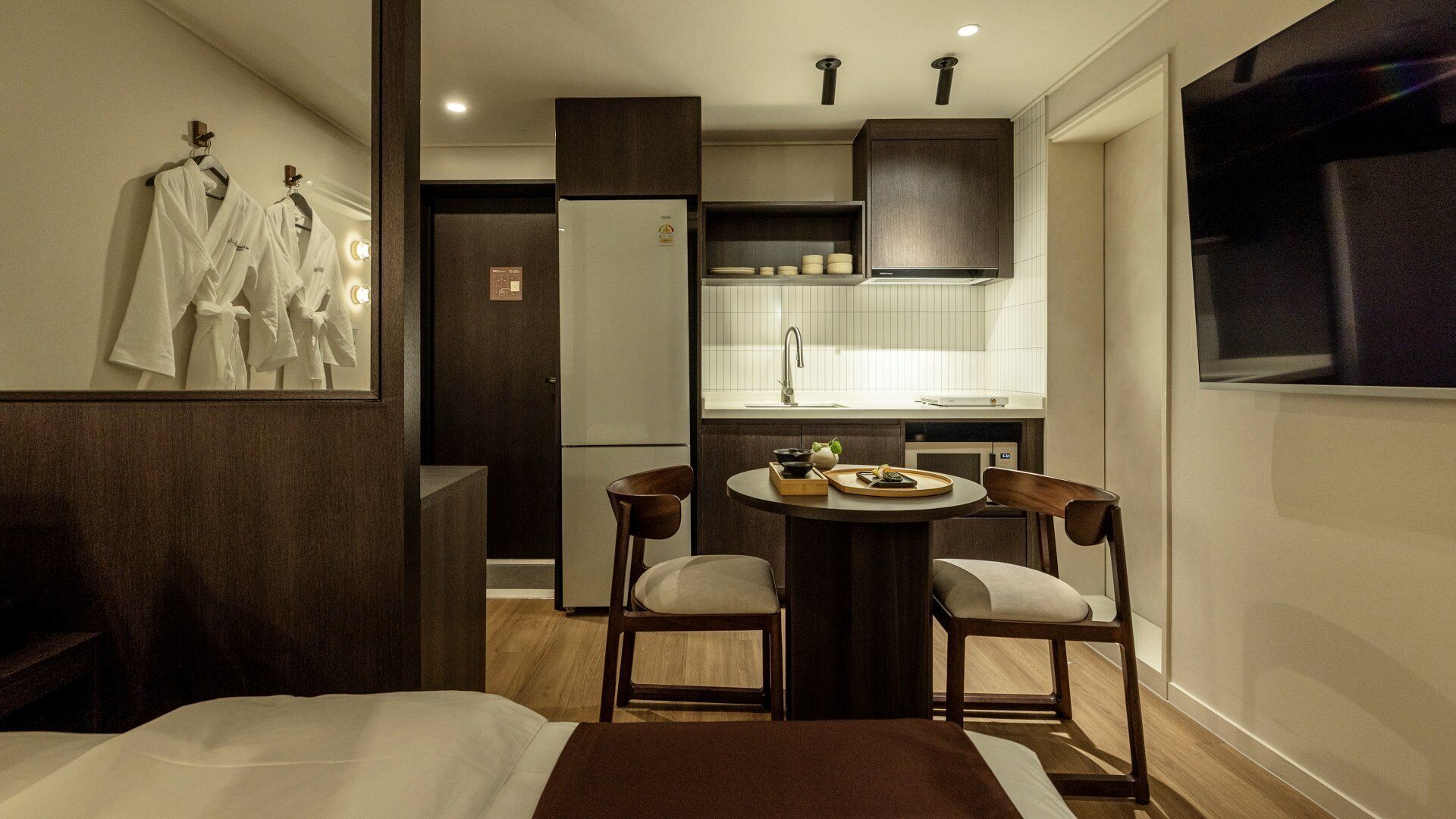 Una habitación de hotel con cama, una pequeña mesa de comedor con sillas, una kitchenette y un televisor montado en la pared.