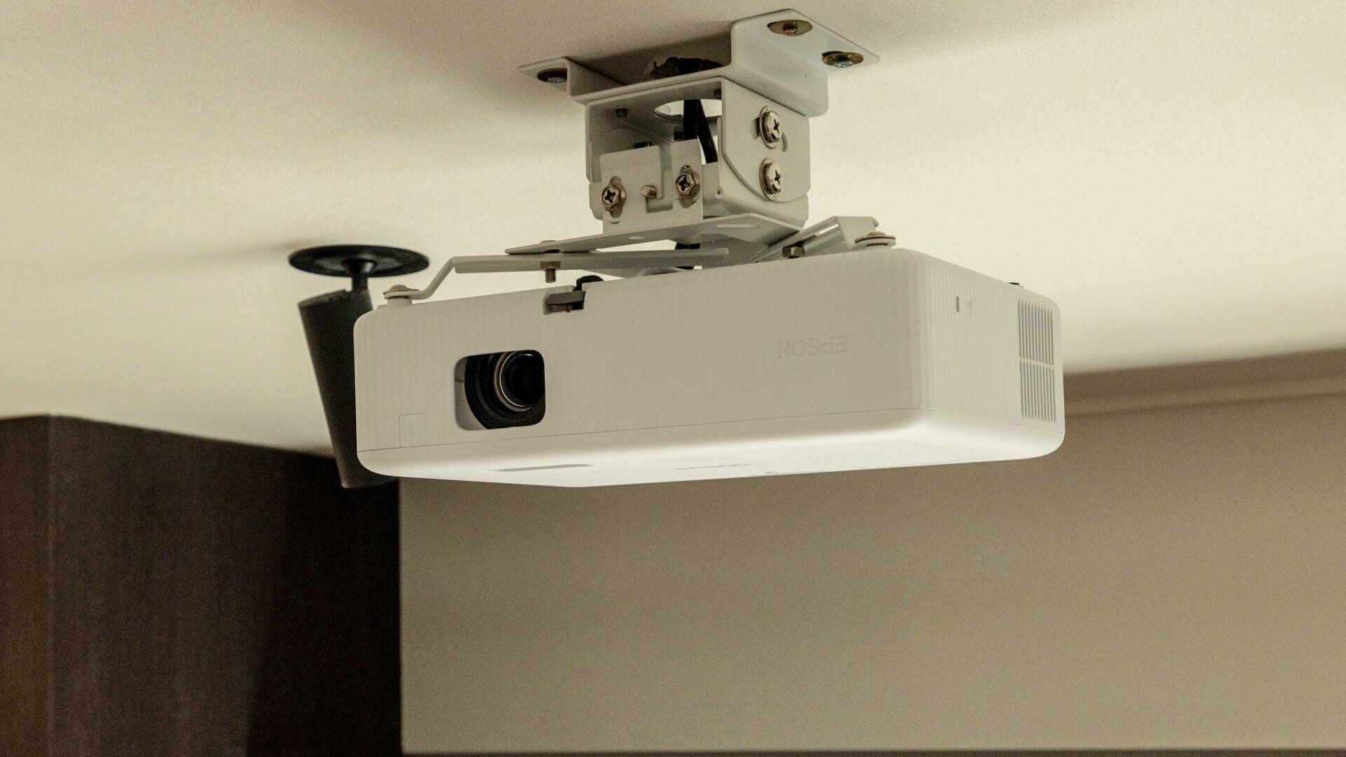 A ceiling-mounted projector with a mounting bracket, a lens, and a ventilation grill.