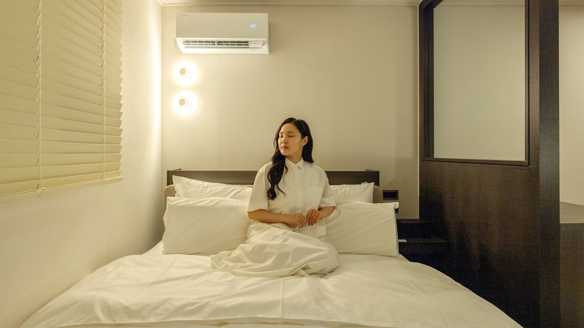 Habitación de hotel con cama, ropa de cama blanca, unidad de aire acondicionado y lámpara fijada a la pared.