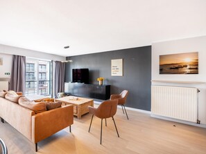Ein modernes Wohnzimmer mit einer Couch, einem Couchtisch und Sesseln, einem Flachbildfernseher, einer dunkel gestrichenen Wand mit einem gerahmten Zitat und einem Fenster mit Vorhängen.