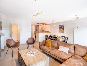 Ein modernes Wohnzimmer mit Sofa, Couchtisch und Sesseln, ein Essbereich mit Tisch und Stühlen sowie eine Küche im Hintergrund.