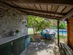A covered patio with a stone wall, a table with chairs, and a view of a swimming pool. - Charming home with garden and private pool. (Edon)
