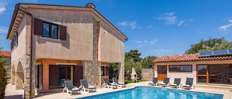 Une maison à deux étages avec une piscine, des murs en pierre et un bâtiment séparé doté d’un toit en tuiles.