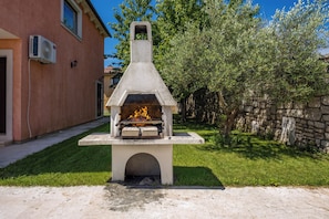 A stone outdoor oven with a fire burning inside, set on a concrete base in a grassy area with a house in the background. - Pool House Suzi in Heart of Istria (Barban)
