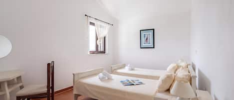 A bedroom with a bed, a chair, a bedside table with a lamp, a framed picture on the wall, and a window with curtains.