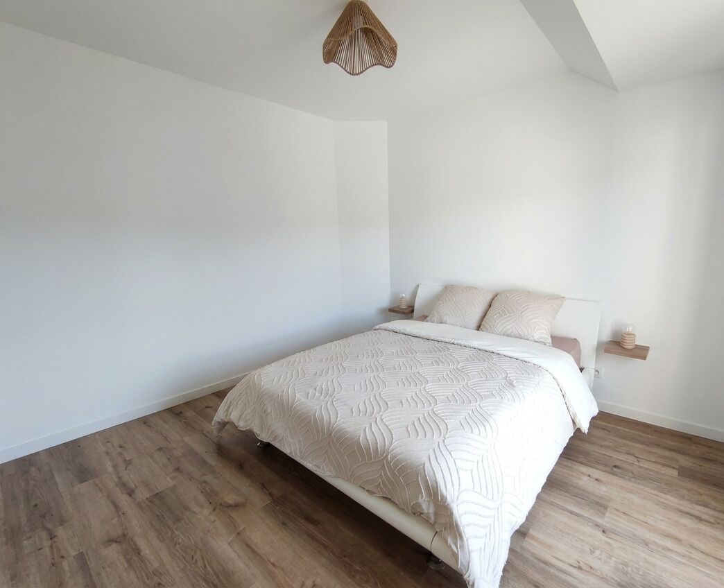 Un dormitorio con cama, suelo de madera, paredes blancas y una lámpara colgante.