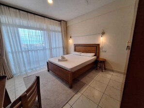 Ein Hotelzimmer mit einem hölzernen Bett, einem kleinen runden Tisch, einem Stuhl und einem Fenster mit durchsichtigen Vorhängen.