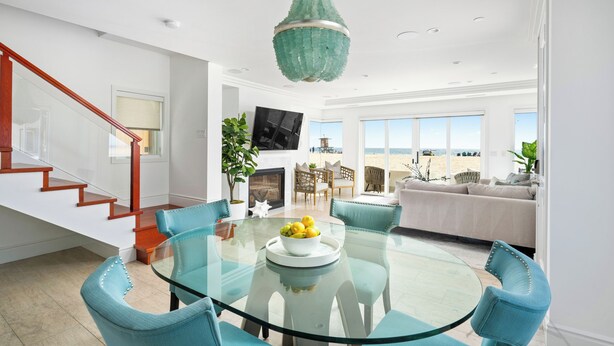 A modern living room with a glass dining table, teal chairs, a fireplace, and a large window overlooking a beach. - OCEANFRONT MODERN LUXURY W/ROOFTOP (Newport Beach)