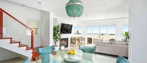 Un salon moderne avec une table à manger en verre, des chaises de couleur turquoise, une cheminée et une grande fenêtre donnant sur une plage.