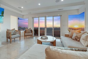Ein geräumiges Wohnzimmer mit einem großen Schiebeglastür, die auf einen Balkon mit Blick auf den Strand und Sonnenuntergang führt.