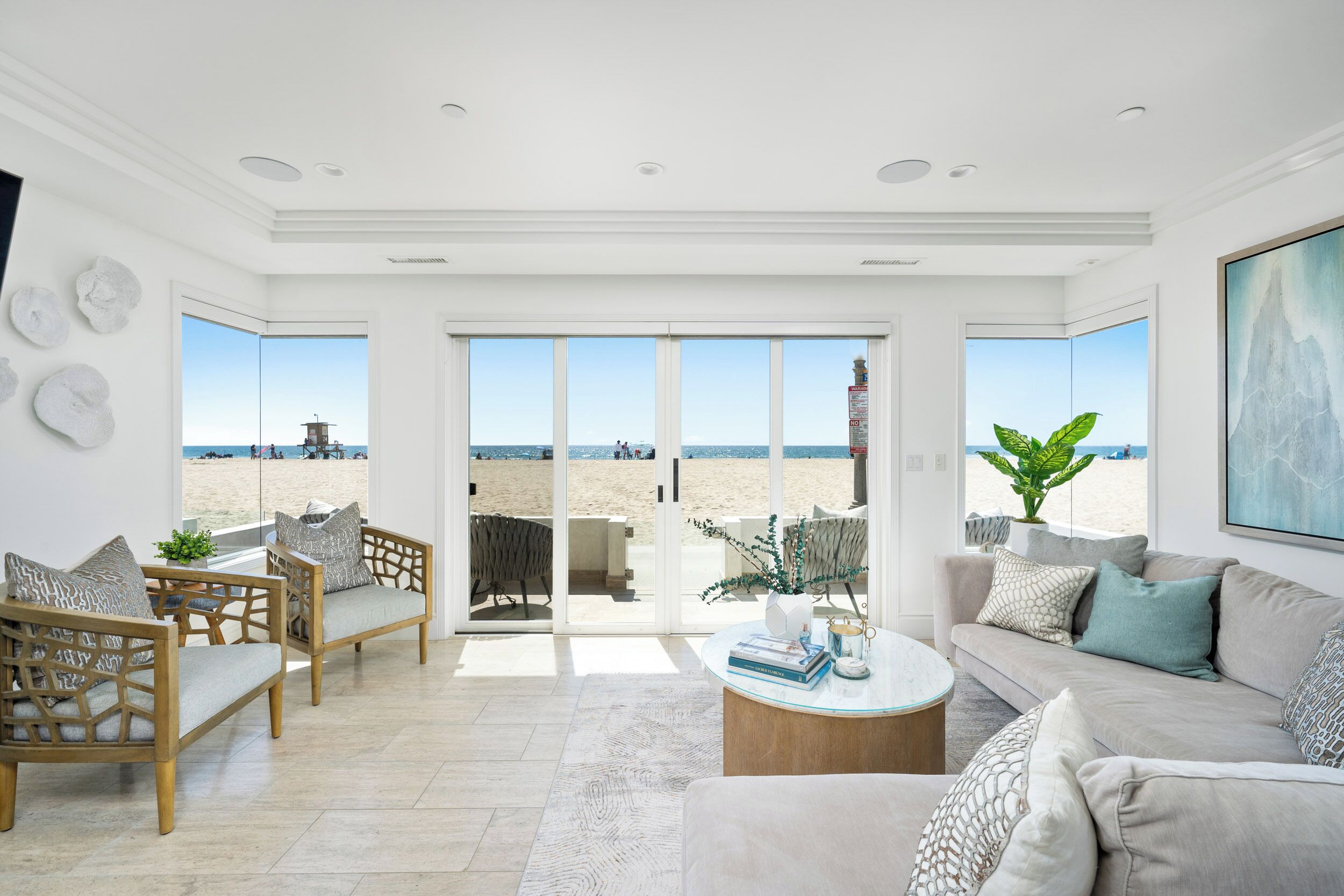 Ein geräumiges Wohnzimmer mit großem Fenster und Blick auf einen Strand, ausgestattet mit einer Couch, Sesseln und einem Couchtisch.