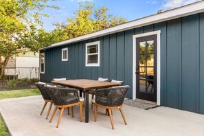 A patio with a wooden table and wicker chairs, a dark blue building, and a door. - Shady Oak: Pool, Hot Tub, Game Room | Sleeps 12 (Fredericksburg)