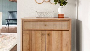 Eine Holzkabine mit einem gewebten Spiegel darüber, eine weiße Vase mit Grünpflanzen und ein kleines, weißes Dekorationsstück auf der Kabine.