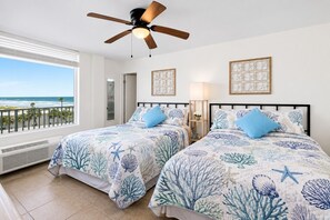 Ein Schlafzimmer mit zwei Betten, einem Deckenventilator, Blick auf den Strand und gerahmten Kunstwerken an der Wand.