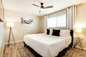 Ein Schlafzimmer mit Bett, Deckenventilator, Fenster mit Vorhängen, Stehlampe und einem Bild an der Wand.