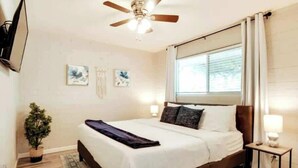 Ein Schlafzimmer mit Bett, Deckenventilator, Fenster mit Vorhängen, Wand-Fernseher und einem kleinen Nachttisch mit Lampe.