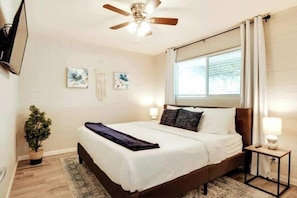 A bedroom with a bed, ceiling fan, window with curtains, wall-mounted TV, and a small bedside table with a lamp. - Casa De Los Caballeros (Scottsdale)