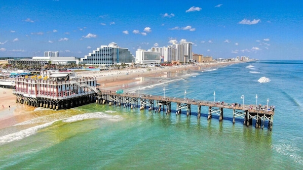 Eine Pier, die sich ins Meer erstreckt, mit einem Gebäude am Ende, einem Strand und einer Stadtansicht im Hintergrund.