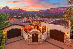 A multi-story house with a stone facade, multiple garage doors, and a view of red rock formations in the background. - Tuscan Red Rock Overlook | Luxury Wellness Villa - 2 Pools, 2 Waterfalls, Spa, Sauna, Fire Features, Massage Chairs & Salt Room! (Sedona)