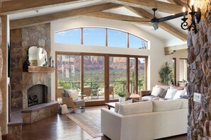 Amplia sala de estar con chimenea de piedra, ventanas grandes que ofrecen vistas a formaciones rocosas rojas y un balcón con mobiliario exterior.
