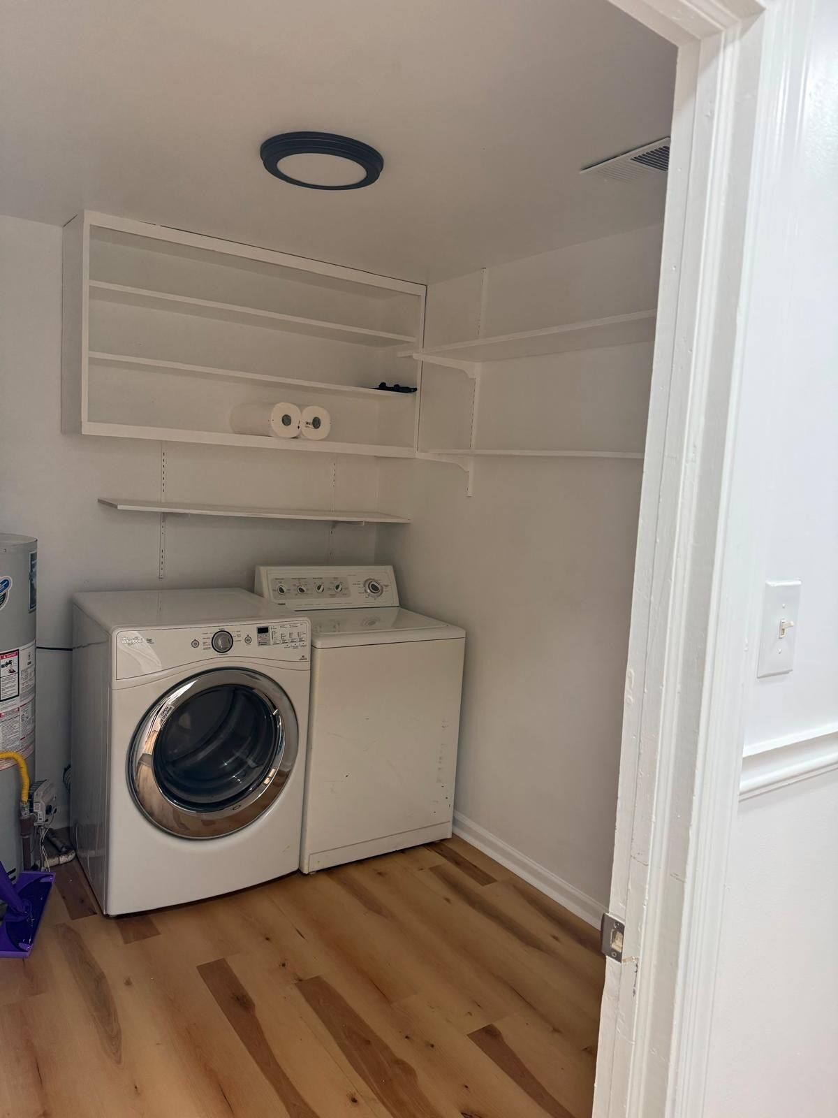 A laundry room with a washing machine, shelves above, and a wooden floor.