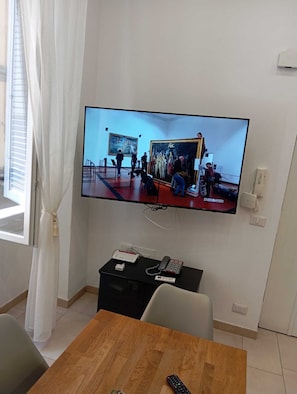 Ein an der Wand befestigtes Fernsehgerät zeigt ein Gemälde, ein Schreibtisch mit Telefon und Fernbedienung sowie ein Fenster mit Vorhängen.