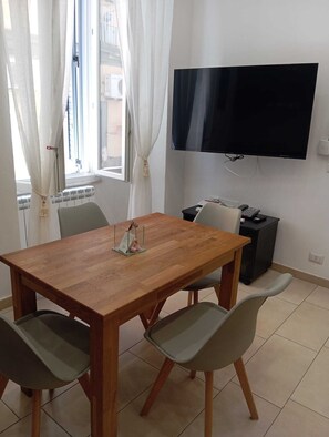 A room with a wooden dining table, four chairs, a TV mounted on the wall, and a small black cabinet.