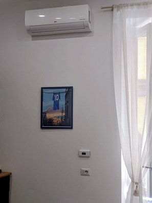 Una habitación con aire acondicionado, un cuadro enmarcado en la pared y una ventana con cortinas translúcidas.