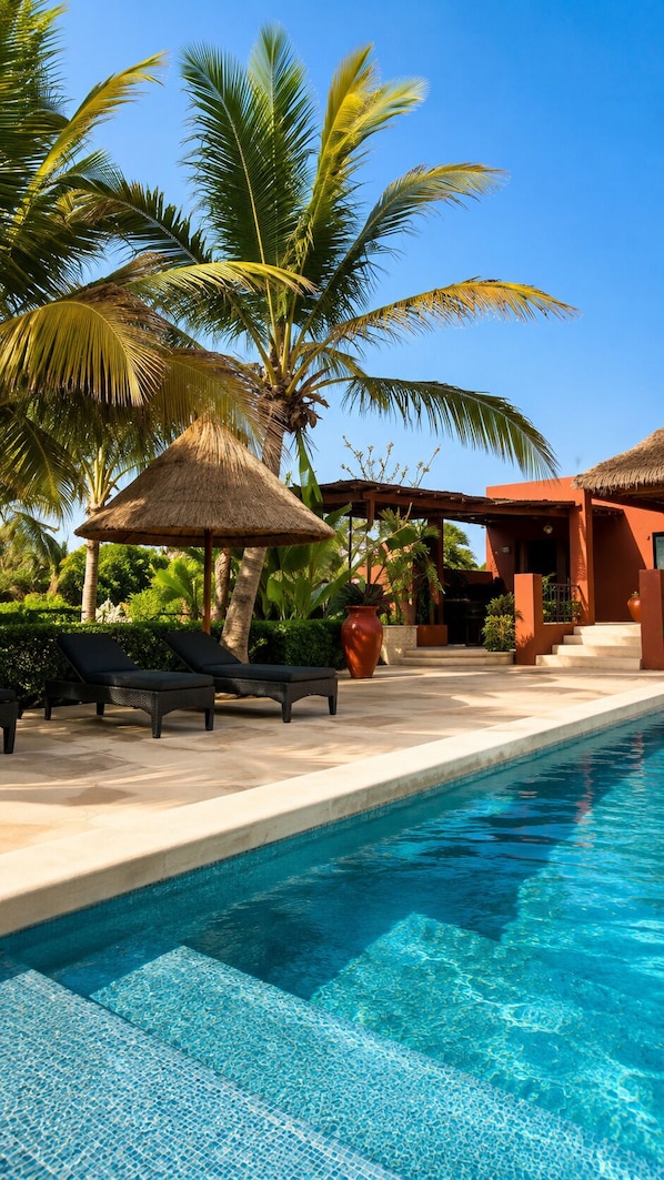 A poolside area with lounge chairs, a thatched umbrella, palm trees, and a building with a red facade.