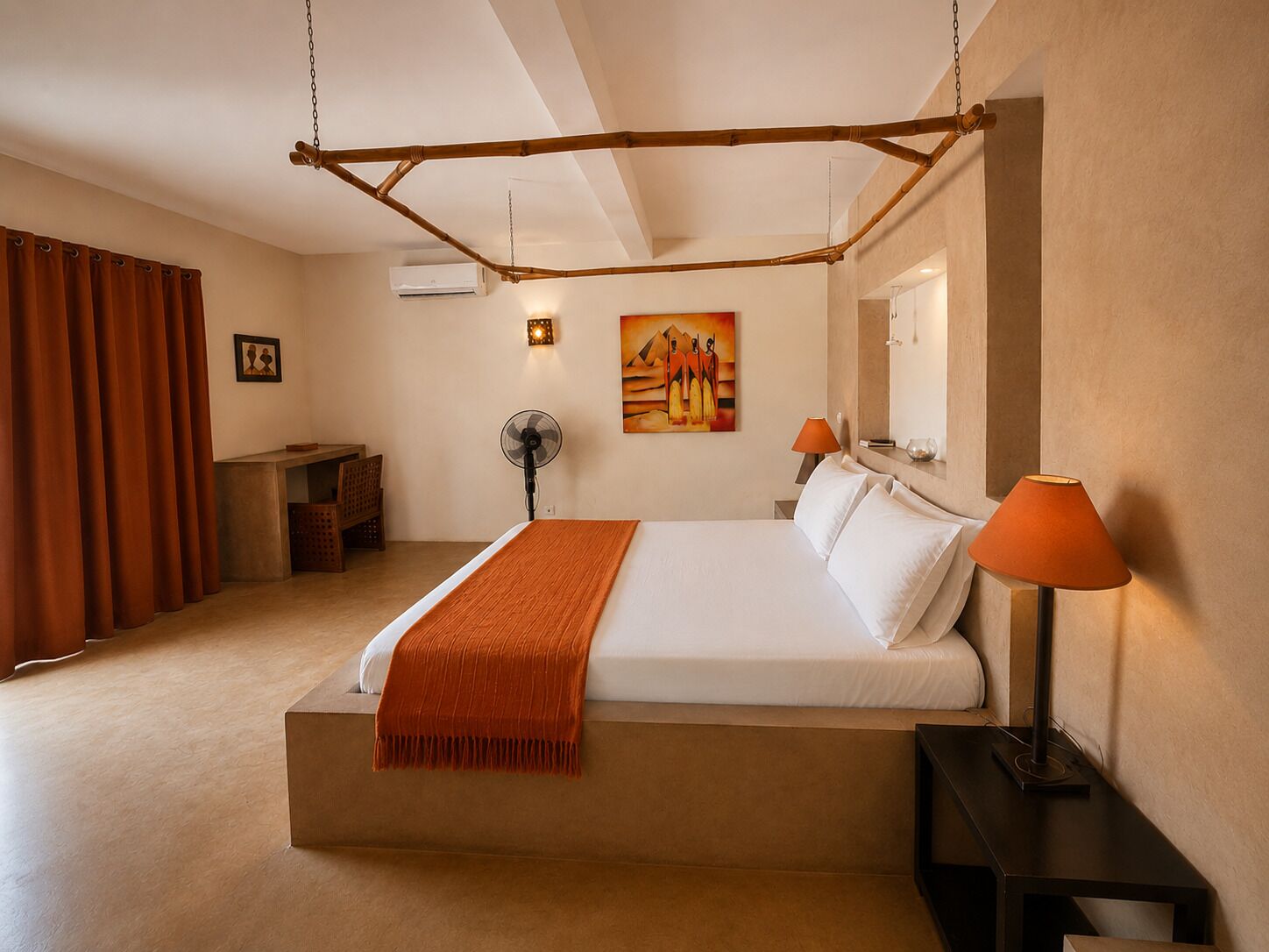 Une chambre d’hôtel avec un lit, une table de chevet, une lampe, un ventilateur, un tableau et une fenêtre avec des rideaux.