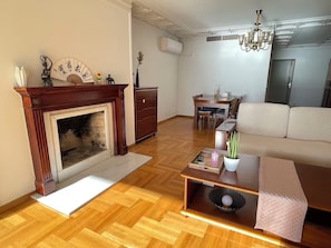 A living room with a fireplace, wooden flooring, a sofa, a dining table with chairs, and a chandelier. - Upscale Sea-View Apartment in Athenian Riviera (Paleo Faliro)