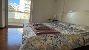 A bedroom with a large bed, a view of buildings through the window, and a small table with a lamp. - Upscale Sea-View Apartment in Athenian Riviera (Paleo Faliro)