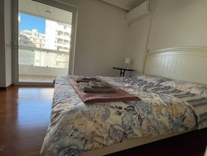 A bedroom with a large bed, a view of buildings through the window, and a small table with a lamp. - Upscale Sea-View Apartment in Athenian Riviera (Paleo Faliro)