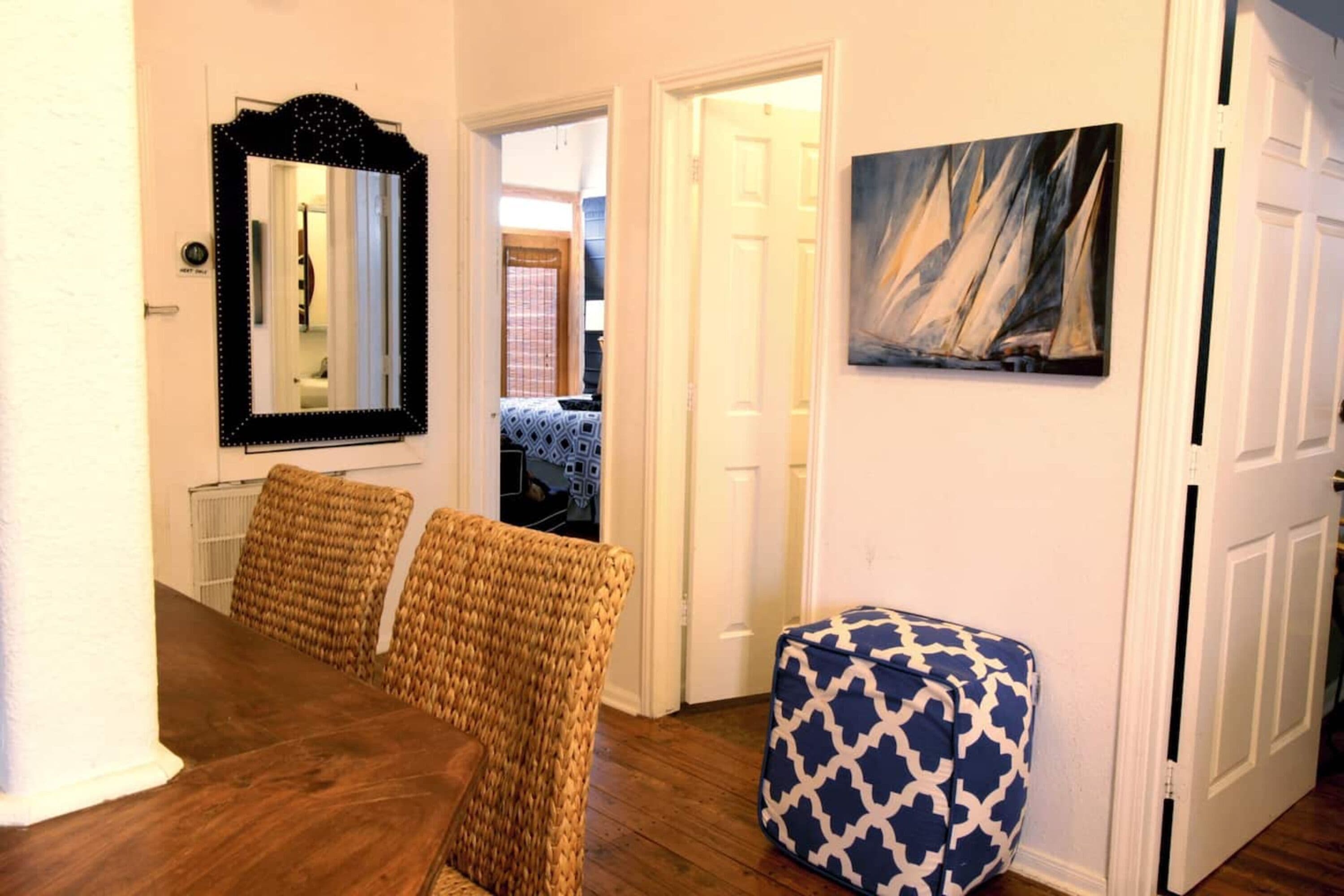 Uma sala de jantar com mesa de madeira e cadeiras de vime, um espelho, um quadro e um banquinho estampado.