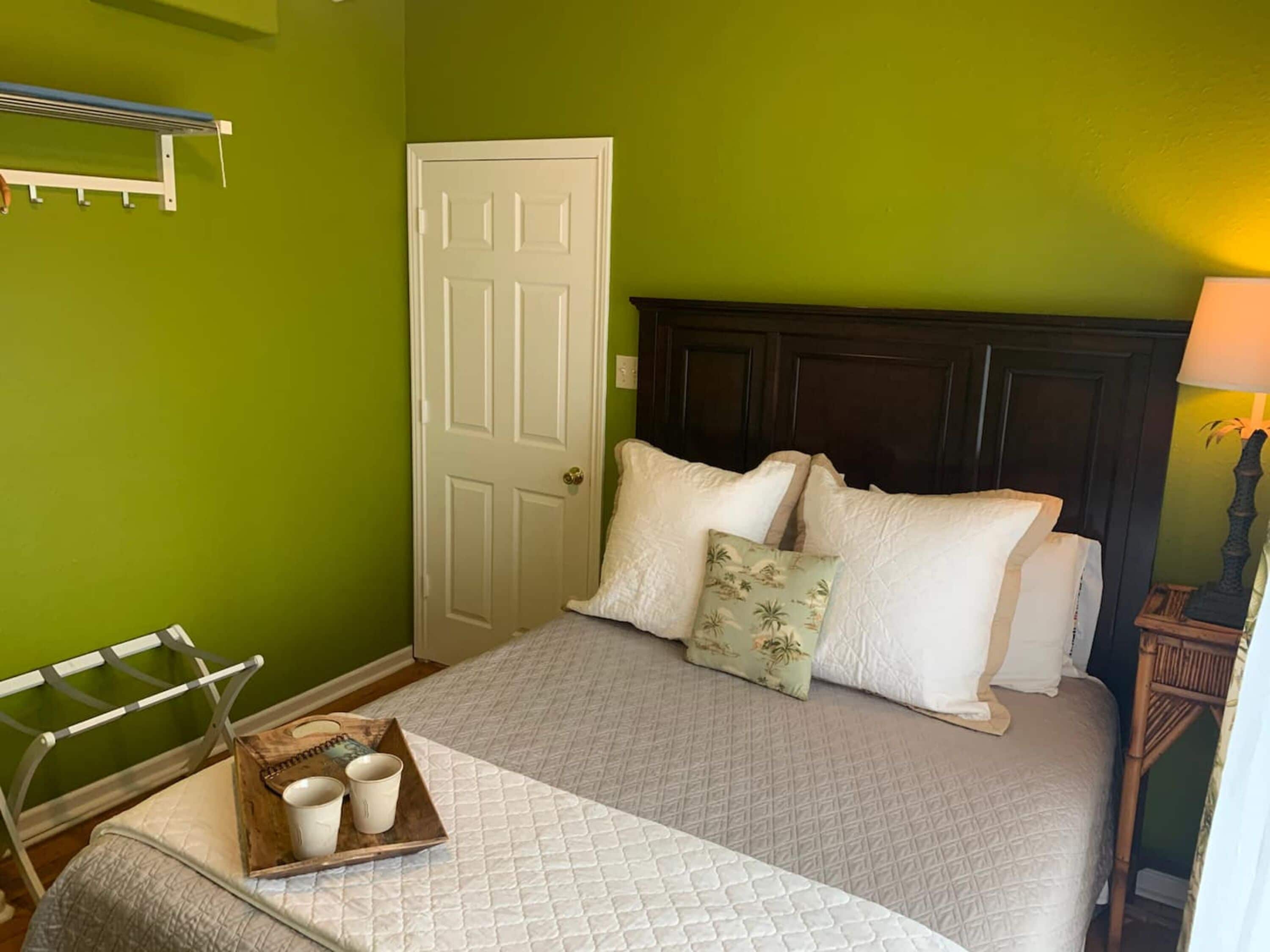 Un lit bien fait, avec des oreillers et un plateau de tasses, une porte blanche et un porte-serviettes fixé au mur, dans une pièce aux murs verts.