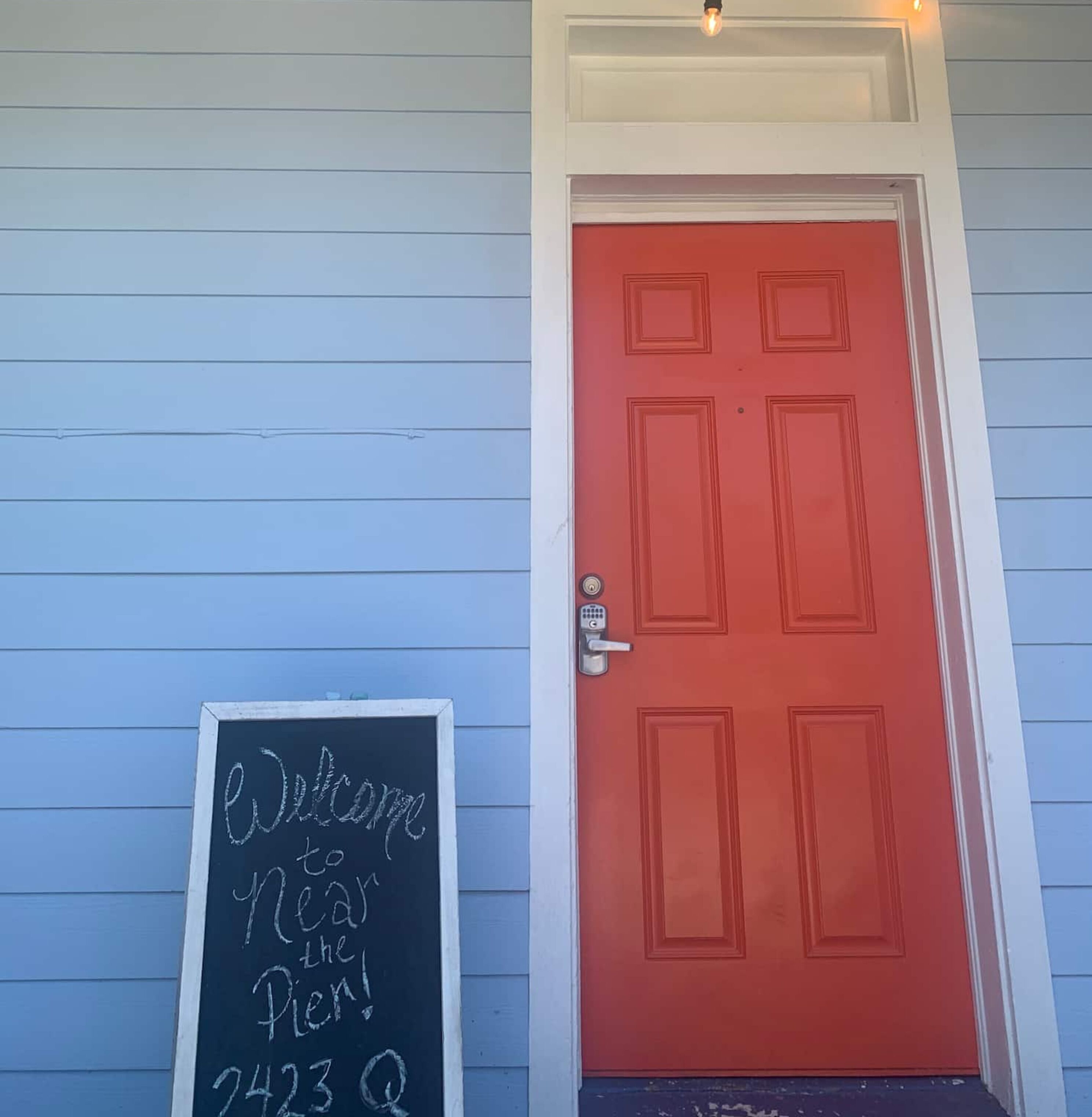 A red door with a keypad lock, a chalkboard sign reading 'Welcome to Pear the Inn,' and a light fixture above the door.