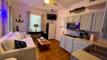 Un espace de vie compact comprenant un canapé, une table basse, un téléviseur, une kitchenette et un ventilateur de plafond.