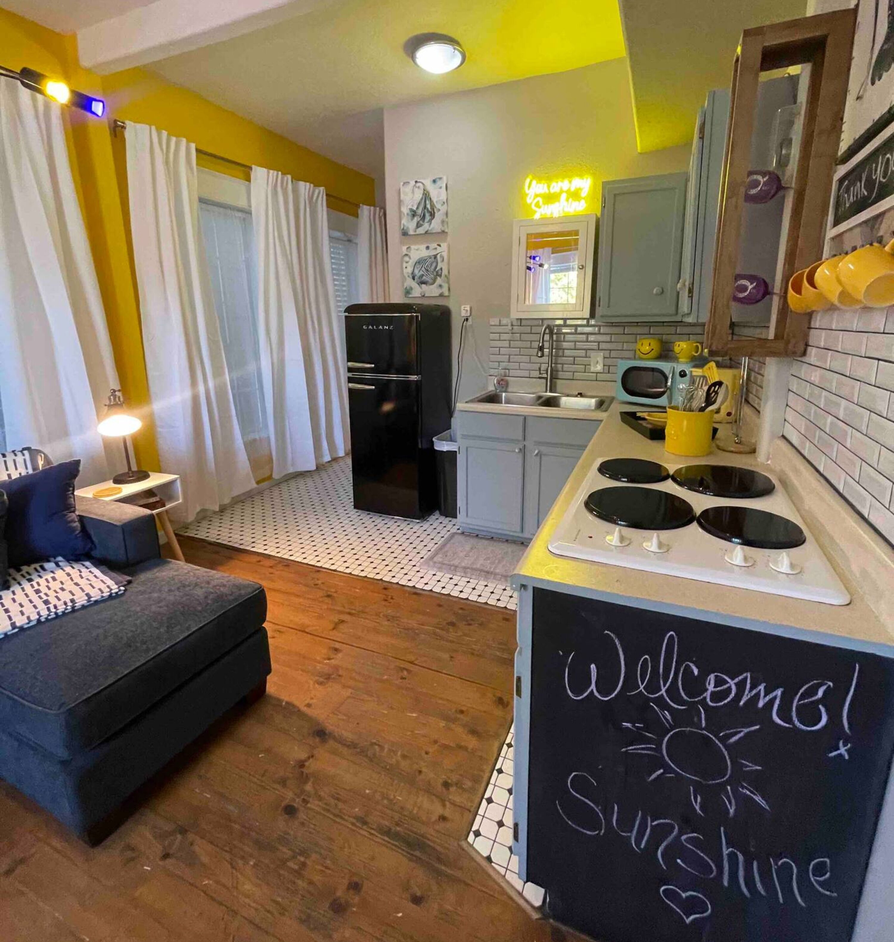 Un espace de vie compact comprenant un coin cuisine, un canapé, un tableau noir sur lequel est écrit « Bienvenue, rayon de soleil », et un panneau néon indiquant « Tu es mon rayon de soleil ».