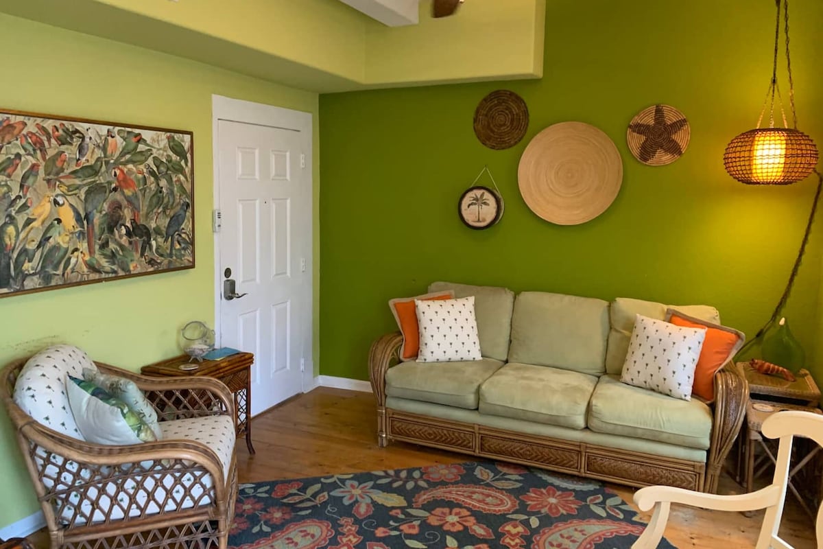A living room with a green wall, a white door, a wicker chair, a sofa with patterned cushions, and a hanging light fixture.