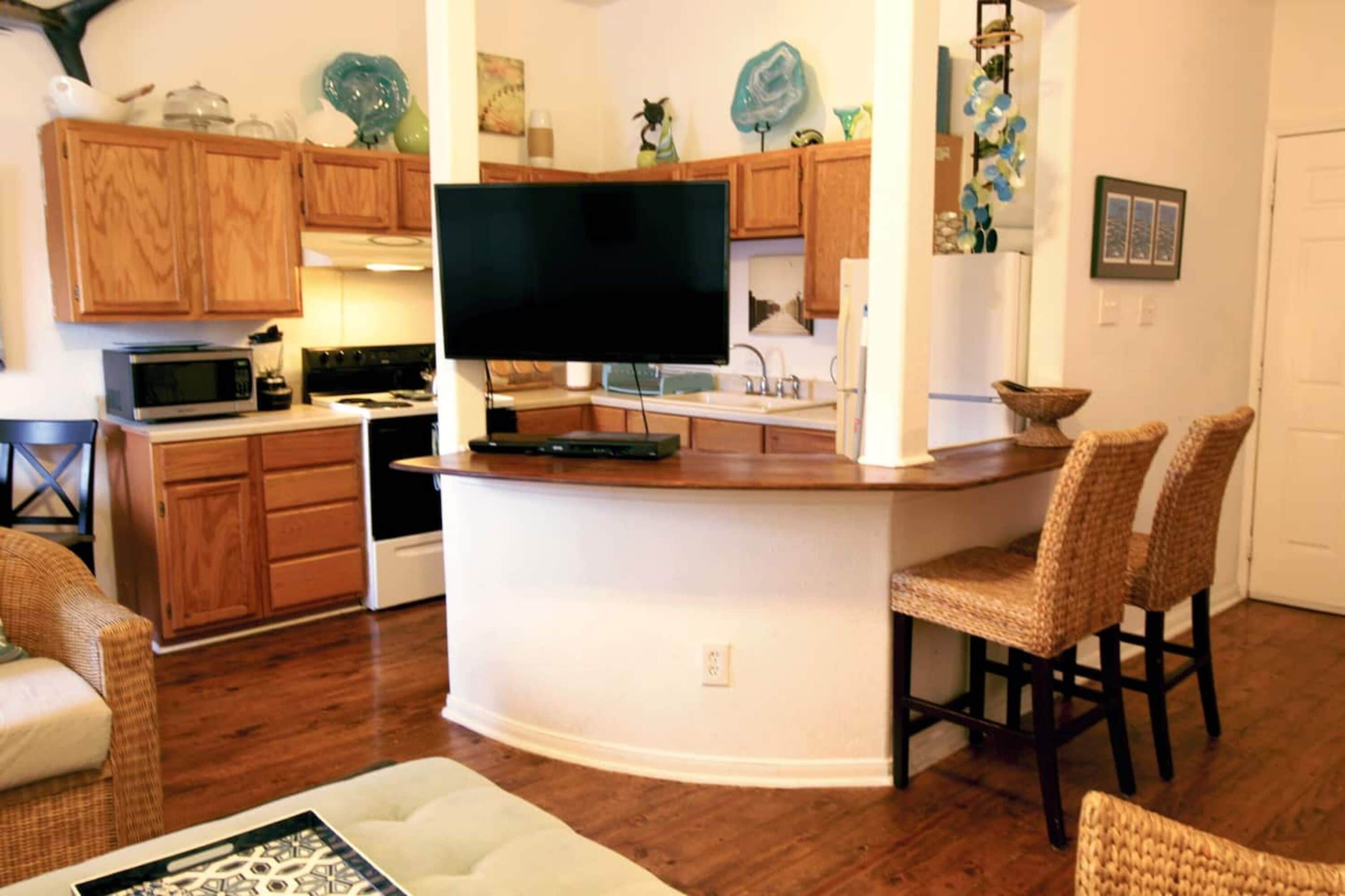 A kitchen with wooden cabinets, a flat-screen TV, a microwave, and a dining area with wicker chairs.